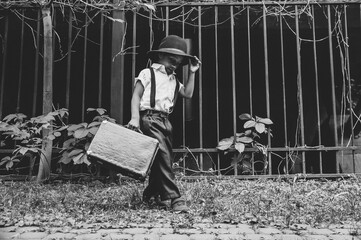 A boy dressed in the style of the 20s dances on the street holding the brim of his hat with a suitcase in his hand