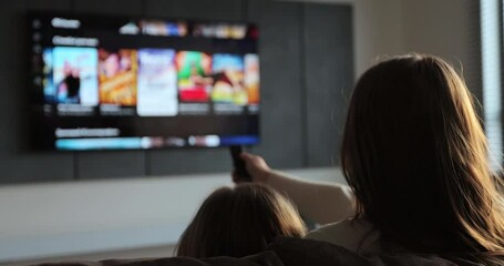 Woman selects tv channels with remote control at living room on sofa. Woman with her little daughter controls TV using a modern remote control.