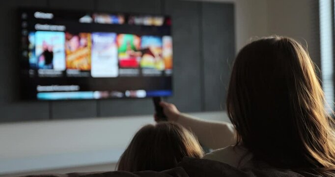 Woman selects tv channels with remote control at living room on sofa. Woman with her little daughter controls TV using a modern remote control.