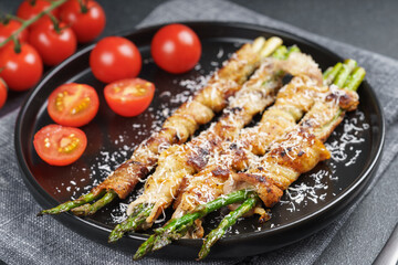 Grilled asparagus wrapped in crispy bacon sprinkled with parmesan cheese served on plate with fresh cherry tomatoes on black background. Gourmet dish, cooking recipe, cuisine, or restaurant menu