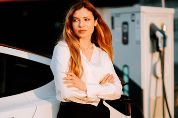 Business woman with red hair charges electric car at charging station in sunny afternoon