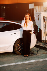 Business woman with red hair charges electric car at charging station during sunset