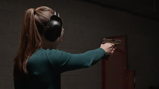 Low angle view of young Caucasian woman wearing goggles and earmuffs shooting handgun at indoor range