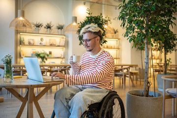 Interested man freelancer in wheelchair working with portable computer in cafe. Focused hipster guy student with disabilities using laptop watches webinar remote study, learning online in coffeeshop.