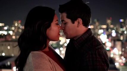 At night atop a rooftop overlooking city lights, a couple stands close, surrounded by the sounds of distant traffic. They lean in and kiss under the stars as wind ruffles their clothes. - Powered by Adobe
