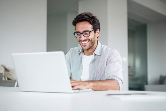 A handsome man in a shirt and glasses is sitting at a white table with a laptop, smiling while working on a digital marketing project in a modern office room with copy space.