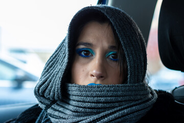 Young woman wearing gray hood and scarf with vivid blue makeup in car