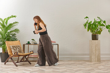 A woman with glasses sits in a woven chair, looking at a phone. She wears a black top, dark pants, and cozy grey slippers.