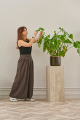 A woman in a black top and dark wide leg pants waters a large plant. The plant sits in a dark pot on a light-colored pedestal.