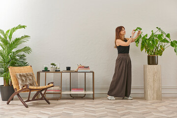 A woman in a black top and dark wide leg pants waters a large plant. The plant sits in a dark pot on a light-colored pedestal.