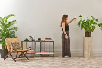 A woman in a black top and dark wide leg pants waters a large plant. The plant sits in a dark pot on a light-colored pedestal.