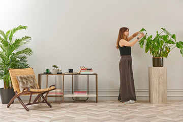 A woman in a black top and dark wide leg pants waters a large plant. The plant sits in a dark pot on a light-colored pedestal.