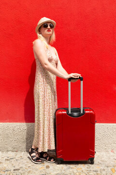 Woman in summer dress travelling with red suitcase in front of a red painted wall