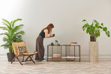 A woman in a black top and dark wide leg pants waters a large plant. The plant sits in a dark pot on a light-colored pedestal.