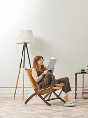 A woman is reading a book at home, looking at the phone and tablet. Decorative interior style and vase of plant.