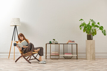 A woman is reading a book at home, looking at the phone and tablet. Decorative interior style and vase of plant.