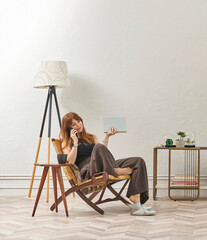 A woman is reading a book at home, looking at the phone and tablet. Decorative interior style and vase of plant.