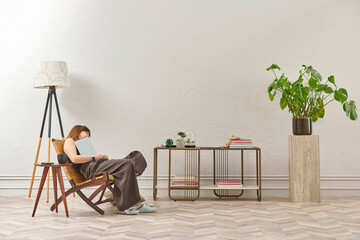 A woman is reading a book at home, looking at the phone and tablet. Decorative interior style and vase of plant.