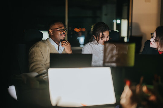 A group of diverse colleagues working late into the night, collaborating on innovative projects. They are engaged in brainstorming to meet deadlines and strategize effectively around a glowing laptop.