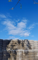 Neatly stacked concrete paving blocks with rough textures under a bright blue sky and scattered clouds, with tree branch shadows adding natural contrast