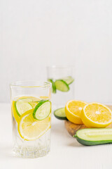 Fototapeta premium Refreshing homemade healthy detox water infused with juicy yellow ripe lemon citrus fruit and green cucumber slices served in drinking glass on white wooden table with copy space and ingredients