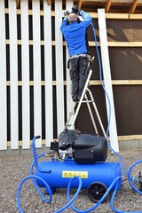 A man stands on a stepladder and holds a pneumatic stapler in his hand - a nail gun for a wooden board, hammering metal nails and staples using a compressor.