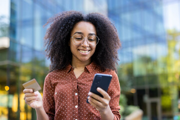 An attractive woman using her phone while holding a credit card in an urban setting.