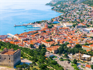 Fototapeta premium Aerial image of the picturesque town of Senj, Croatia, showing the Nehaj Fortress, old buildings with red-tiled roofs, and a beautiful blue coastline with a harbor full of boats