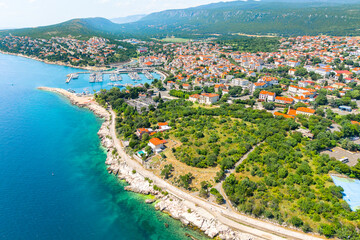 Stunning aerial panorama of Novi Vinodolski, showing the coastal town with red-roofed houses, marina, rocky beaches, and lush green hills surrounding the blue Adriatic Sea