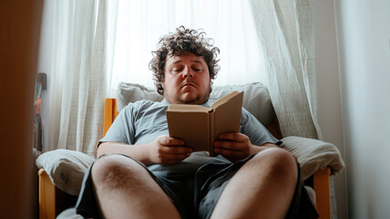 Young caucasian male relaxing on a chair while reading a book, engaged in leisure activity in a cozy room with soft lighting and curtains. Overweight teen boy