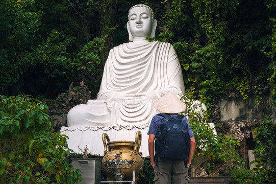 A male traveler in a Vietnamese hat at the white marble Buddha statue on the Marble Mountains in Da Nang.