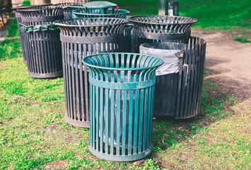 Cylindrical outdoor trash cans with vertical slats, placed on a grassy area. Cans contain plastic...