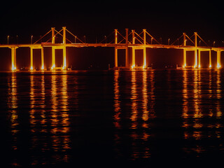 Illuminated Bridge In Hong Kong At Night: Glowing Golden Lights Reflecting On Calm Water Surface Beneath A Modern Suspension Bridge In A Coastal Urban Landscape