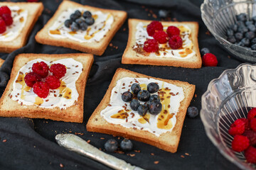 Flat lay of toasted white bread topped with labneh, fresh raspberries, blueberries, maple syrup, and flax seeds. Sweet breakfast or brunch idea with natural light and food styling