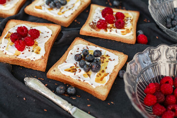 Flat lay of toasted white bread topped with labneh, fresh raspberries, blueberries, maple syrup, and flax seeds. Sweet breakfast or brunch idea with natural light and food styling