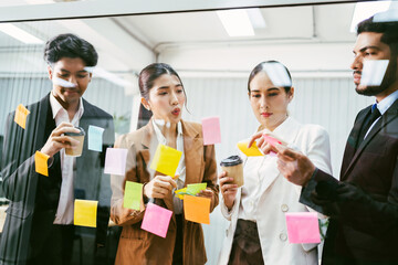Multicultural office team collaborates in a brainstorming session, placing sticky notes on a glass wall. Business planning, creativity, and teamwork in a modern work environment with coffee in hand.
