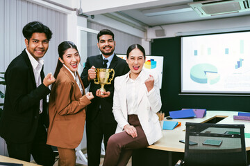 Diverse business team celebrates corporate success with a trophy in a modern office. Charts on screen suggest teamwork, achievement, and strategic consulting in a thriving work environment.