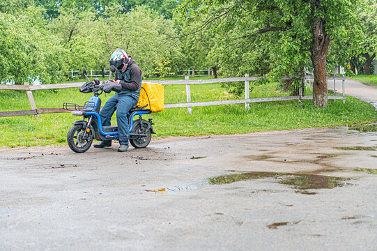 Person on blue electric motorcycle, wearing helmet, adjusting handlebars, yellow cargo box, wet ground, puddles, ruralsuburban setting, trees nearby, daytime under overcast conditions, candid photog