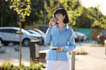 Work life balance. Adult confident elegant businesswoman talks on smartphone, holds cup of take away coffee outdoors on city street. Business concept, urban people, city lifestyle working people.
