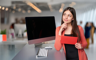 Young white woman in office holding red envelope and looking thoughtful beside desktop computer. Concept of career goals, planning future decisions, and professional development.