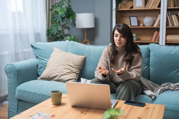 Young woman attending online therapy session from her living room, expressing emotion while talking...