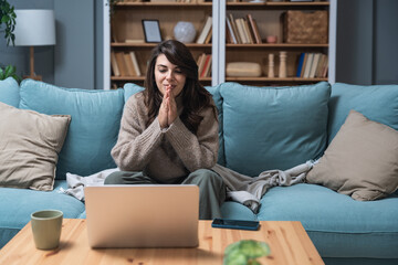 Young woman attending online therapy session from her living room, expressing emotion while talking...