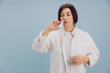 Middle aged woman using nasal spray for treatment on blue background