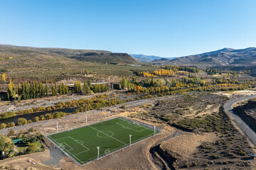 Aerial view of soccer field nestled in a valley with autumn foliage next to Nahueve river and distant hills. Villa del Nahueve, Neuquen, Argentina
