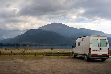 White van motorhome parked near a Lake Moquehue with mountains in the background under a cloudy sky
