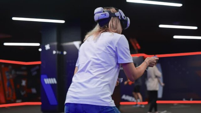 Young woman wearing VR headset with motion controllers playing an immersive shooter game with friends at a specialized arena. Dynamic virtual reality experience in neon-lit dark room.