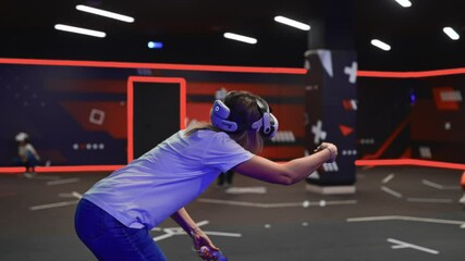 Young woman wearing VR headset with motion controllers playing an immersive shooter game with friends at a specialized arena. Dynamic virtual reality experience in neon-lit dark room. - Powered by Adobe