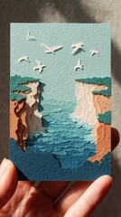 Resilient Coastline Papercut Art - Hand holding card of coastal cliff with waves, seagulls, symbolizing change and strength
