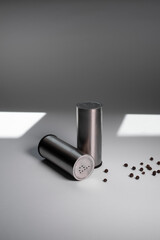 Metal pepper shaker and salt shaker, peppercorns next to a white background with light from the sun