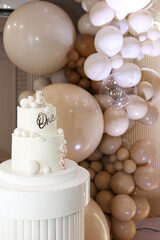 Elegant white birthday cake decorated with bear toppers and “One” sign, set against a stylish backdrop of beige and white balloons for a first birthday celebration.